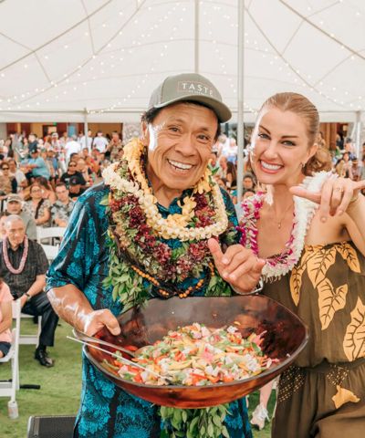 Two people in tropical outfits pose smiling and making &ldquo;shaka&rdquo; signs by a large laulau-style dish at a festive outdoor event under string lights.