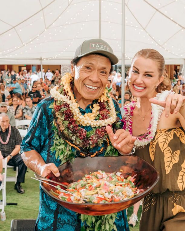 Two people in tropical outfits pose smiling and making &ldquo;shaka&rdquo; signs by a large laulau-style dish at a festive outdoor event under string lights.