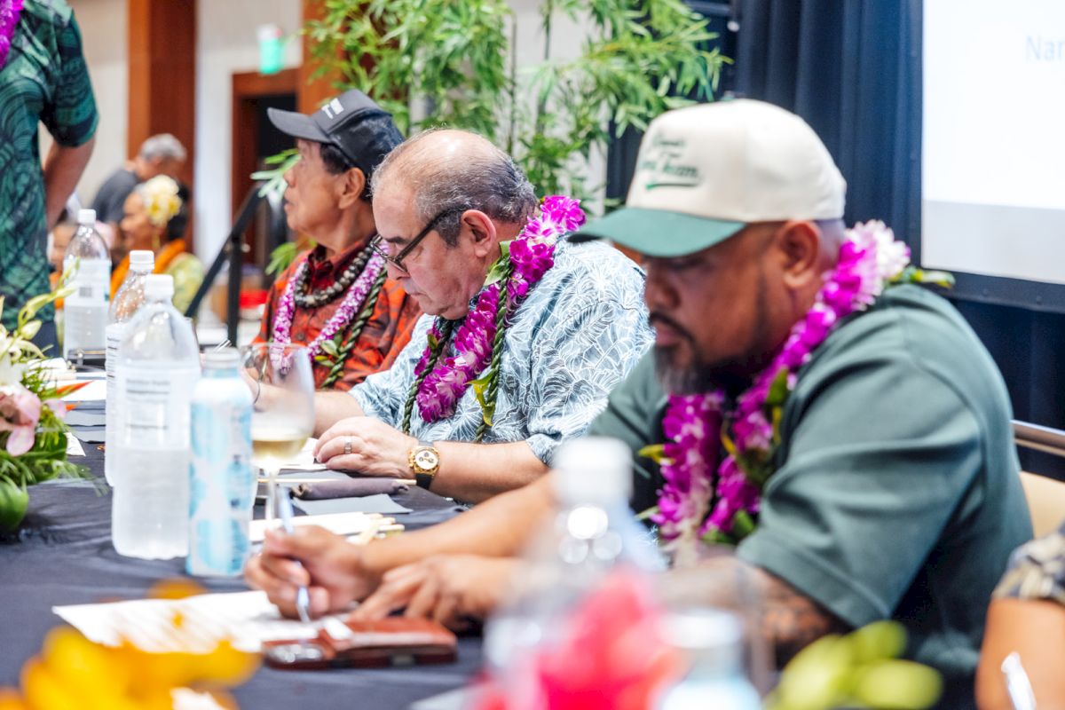 Several men in Hawaiian shirts and purple leis sit at a table, one wearing glasses writing while others listen.