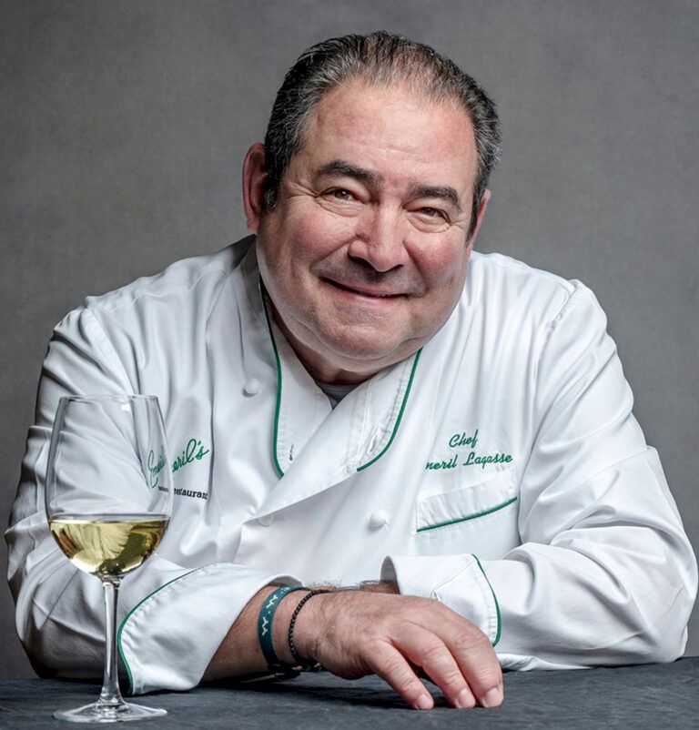 A smiling chef in a white coat with green trim sits at a table with a wine glass in front of him.