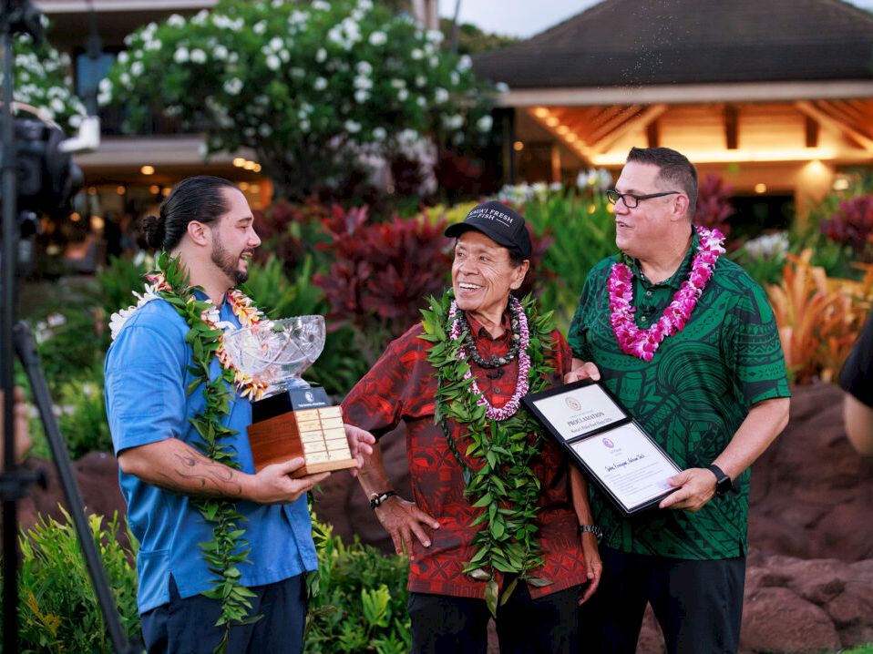 Three people in colorful shirts and leis present awards outdoors at a garden event, holding certificates and smiling.