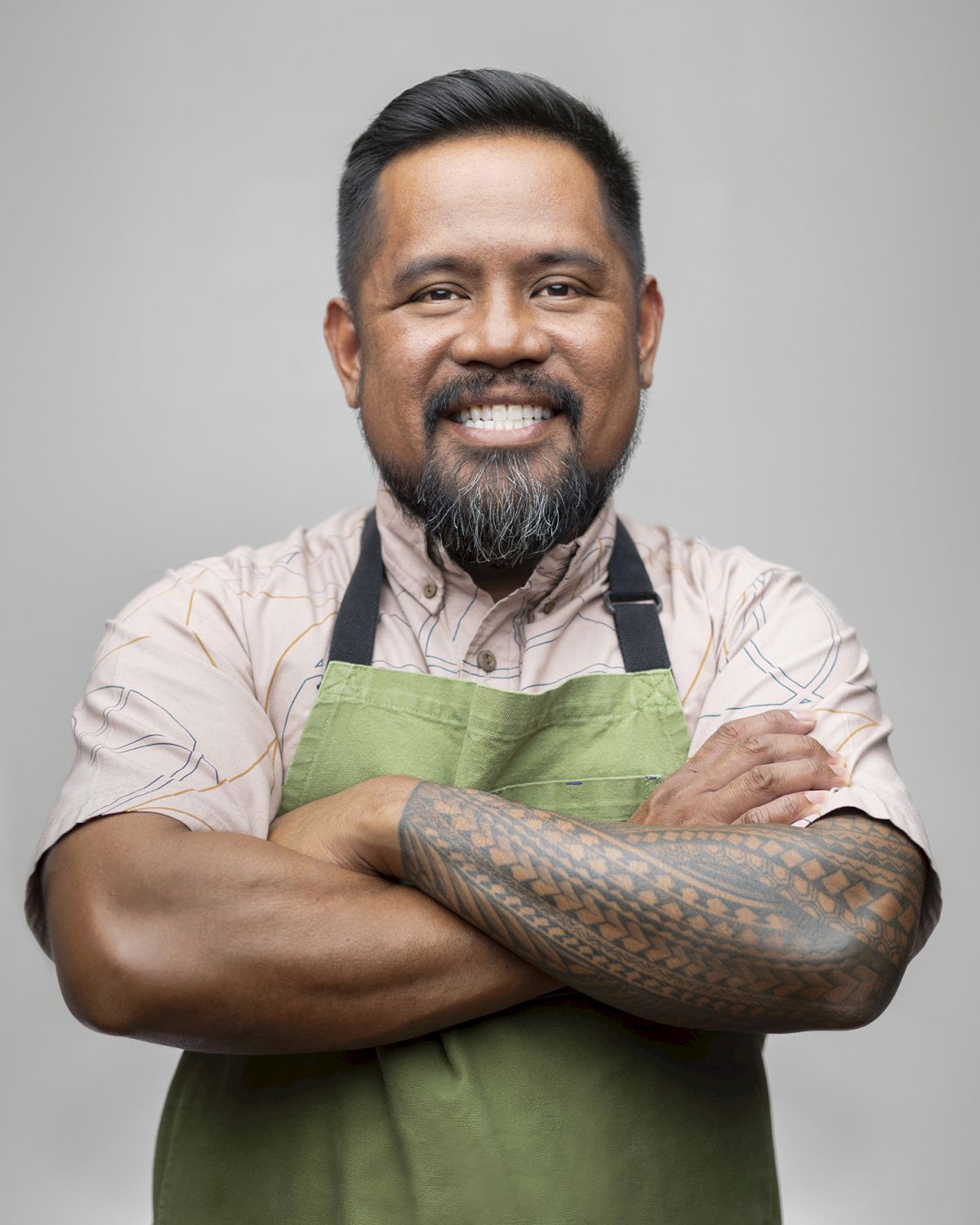 A smiling chef in a green apron poses with arms crossed against a plain gray background.