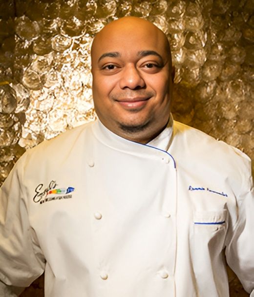 A smiling bald chef in a white double-breasted jacket poses in front of a textured golden wall.