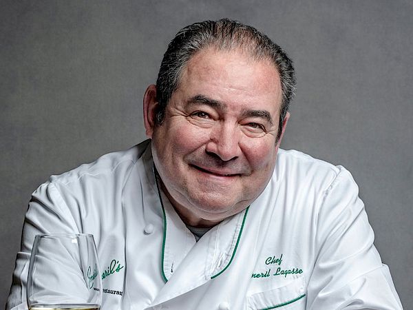 A chef in a white coat poses at a table with a glass of white wine against a plain gray background.