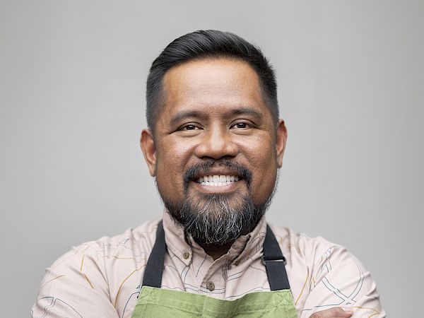A smiling man wearing a green apron with tattooed arms, posing with his arms crossed against a plain light background.