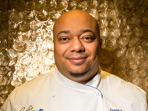 A smiling chef in a white double-breasted jacket stands before a gold, textured backdrop.