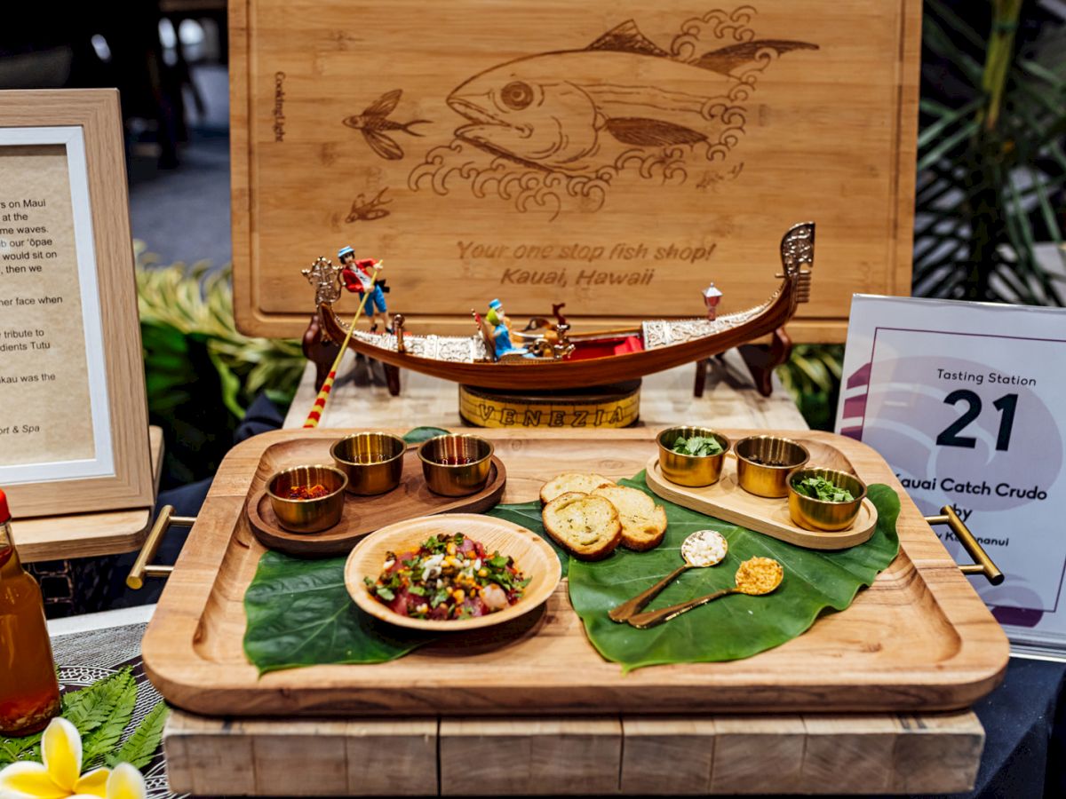 A Hawaiian-themed fish tasting station with a wooden board, small gold bowls, breads, greens, and signs reading Tasting Station 21.