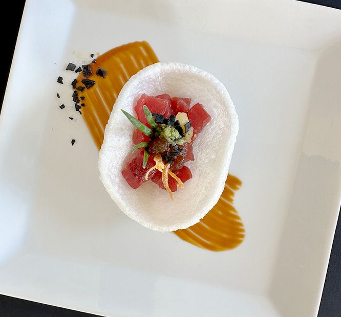 A plated bite of seafood ceviche: red tuna atop a white round with herbs, orange sauce, and black crisp bits.