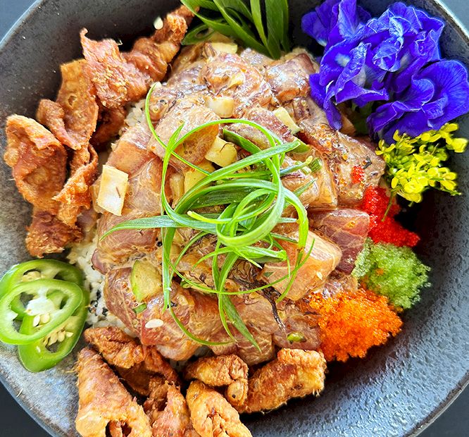 A plated meal with crispy pork, shredded greens, purple edible flowers, and garnishes in a bowl.