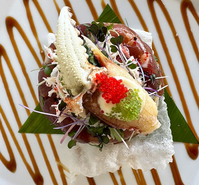 A gourmet seafood tartare on white foam, topped with microgreens, edible flowers, and red/green caviar, on a striped plate.