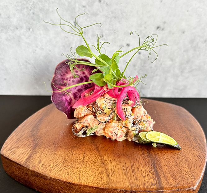 A plated dish with shrimp and beet slices, topped with microgreens and a lime wedge on a wooden round.