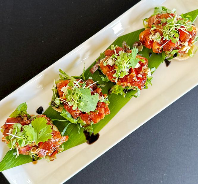 A long white platter holds bites of cured meat on banana leaves, topped with microgreens and herbs, ready to eat.