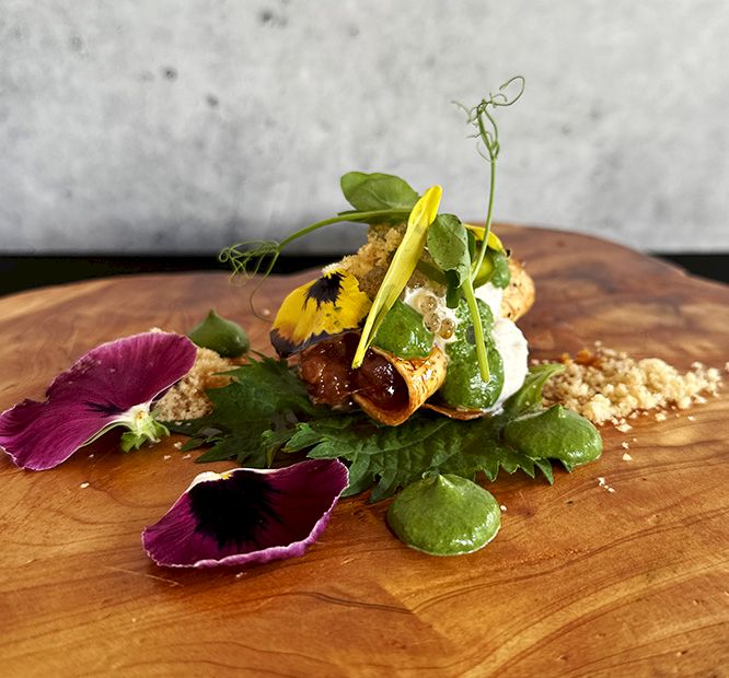 A plated gourmet dish with greens, edible flowers/petals, mushrooms, and a yellow garnish on a wooden board.