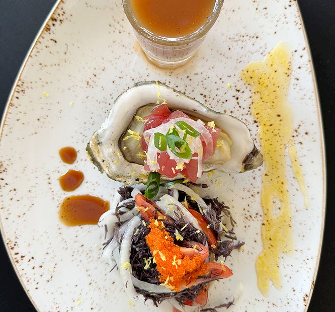 A plated seafood ceviche/oyster topped with diced fish, greens, orange roe, and a dipping sauce, on a white dish.