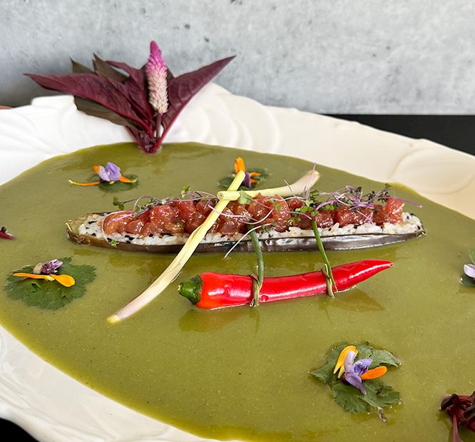 A plated gourmet dish: white fish topped with red salsa, chilies, microgreens, and edible flowers on green puree.
