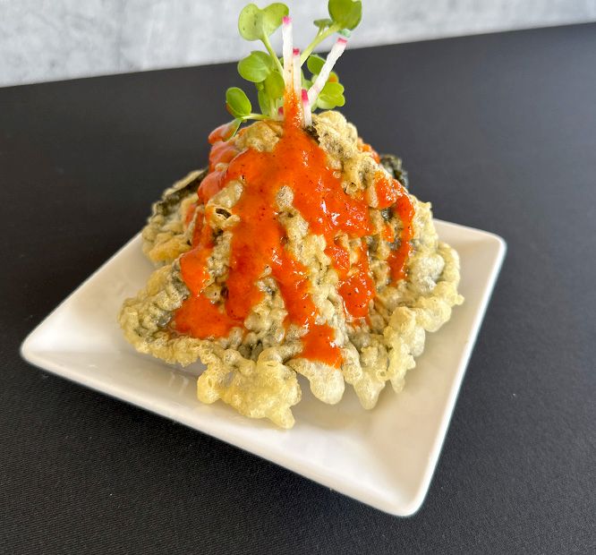 A plated crispy, golden cauliflower topped with bright red sauce, creamy crumble, and tiny green sprouts, ready to eat.