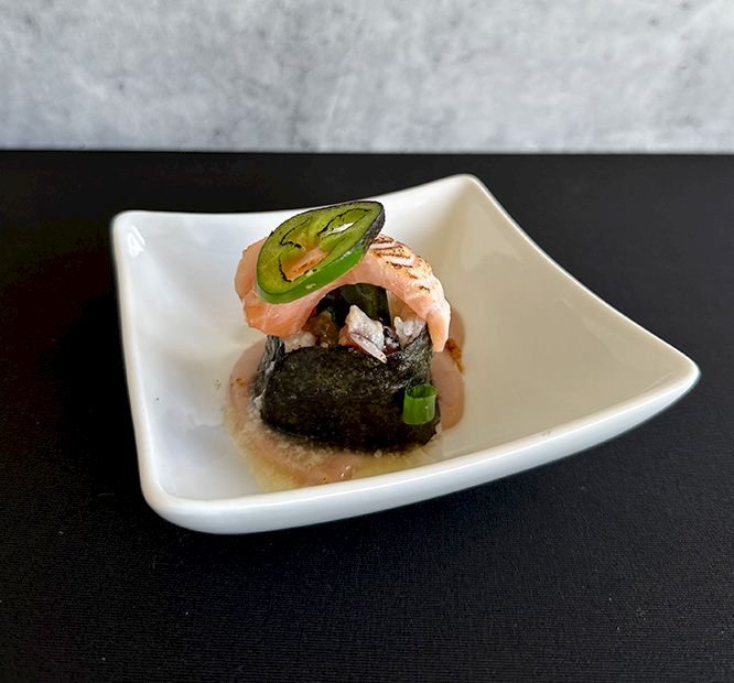 A small plated sushi/nigiri with shrimp, a slice of green avocado, and a dark seaweed topping on a light sauce.