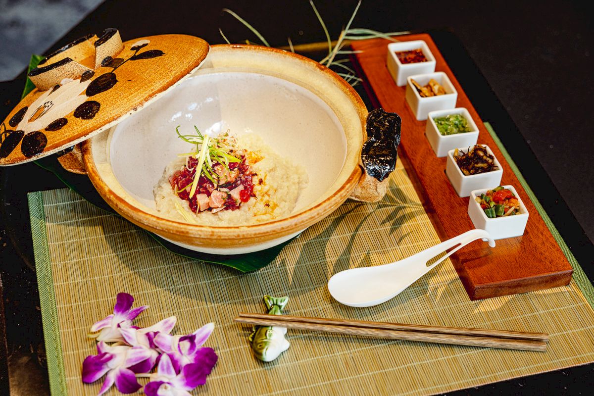 A stone pot of rice porridge topped with herbs and seafood, with small side dishes of sauces on a bamboo mat.