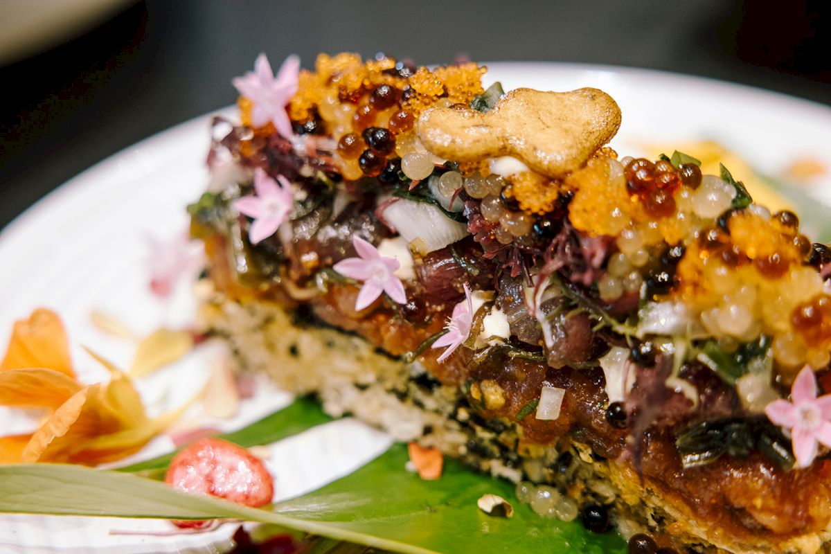 Close-up of a gourmet dish: crunchy meat/p&acirc;t&eacute; with herbs, edible pink flowers, and sauce dots on a plate.