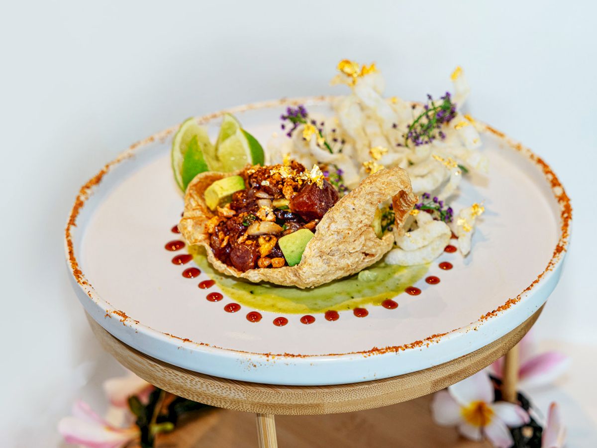 A plated dish with meat, green herb sauce, lime wedges, and purple flower garnish, finished with decorative bits on top.