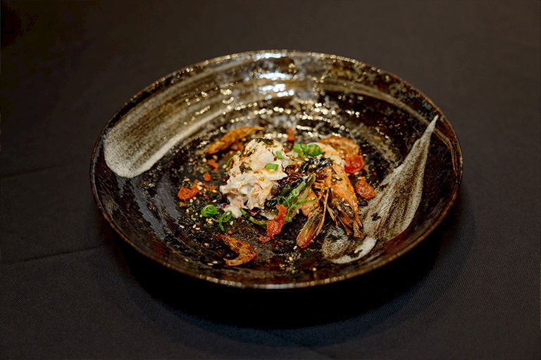 A bowl of noodle soup topped with shrimp, greens, herbs, and diced vegetables in a dark, savory broth.
