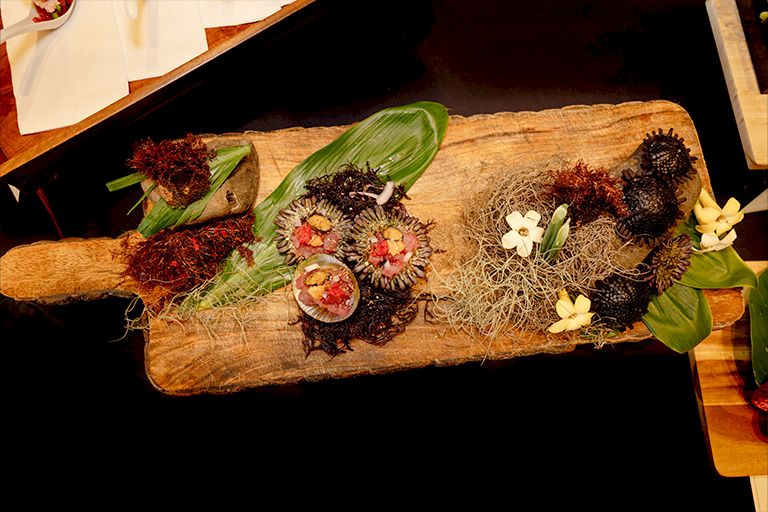 A wooden platter of uni sea urchin roe with seaweed, herbs, and shellfish garnishes over straw and greens.