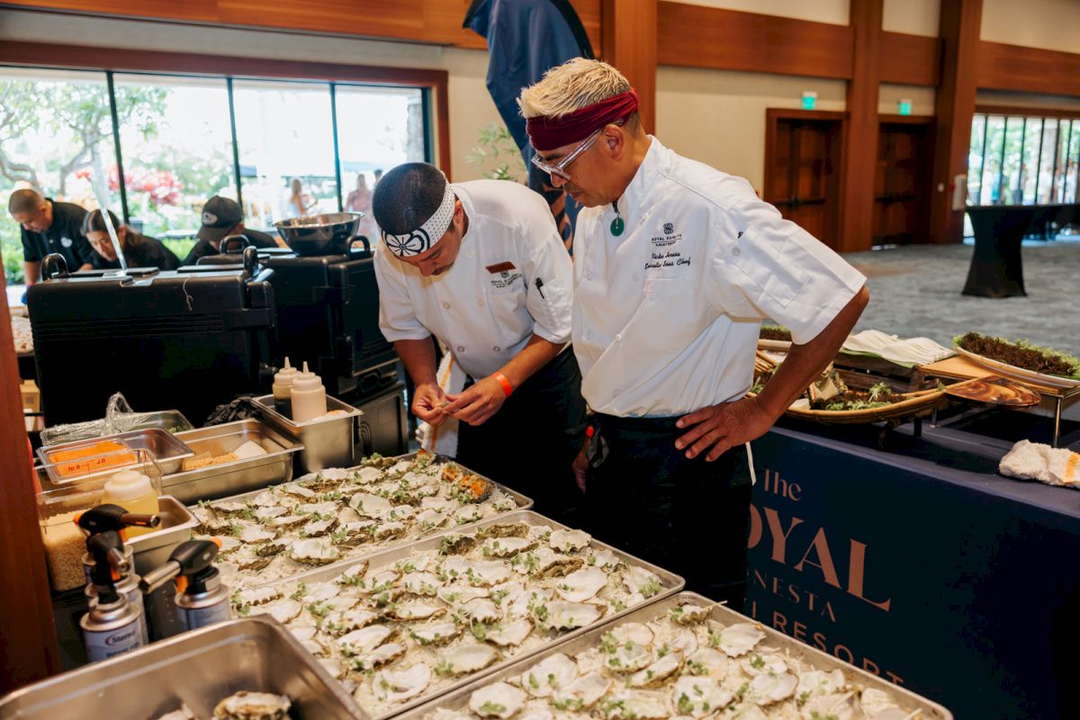 Two chefs in white coats garnish trays of food at a buffet-style cook station, preparing dishes for guests.