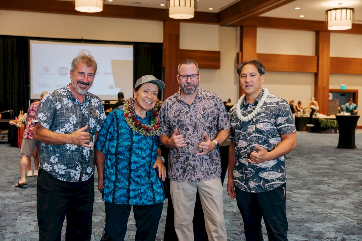 Four men in Hawaiian shirts pose together at an indoor event, smiling in front of a presentation screen.